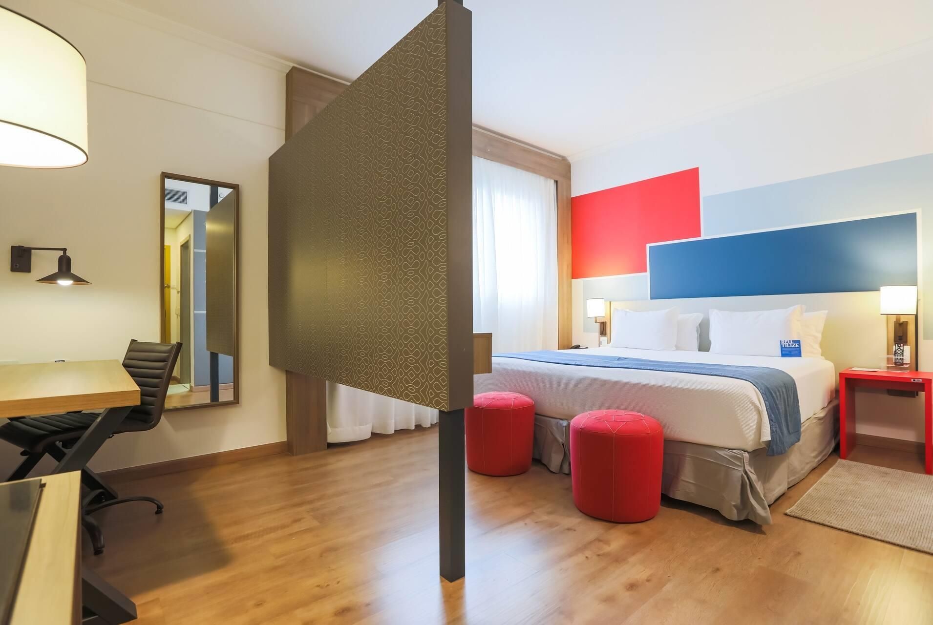 Een hotelkamer met een bed, een bureau, een stoel en een rode poef.