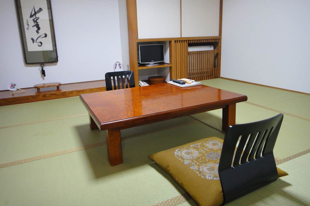Une pièce traditionnelle japonaise avec un sol recouvert de tatamis, une table en bois, des tapis de tatami, une chaise noire, un coussin, un téléviseur et une étagère sur laquelle se trouve un clavier.