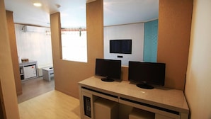 Habitació moderna amb escriptori, dos monitors, televisió i una petita cuineta.