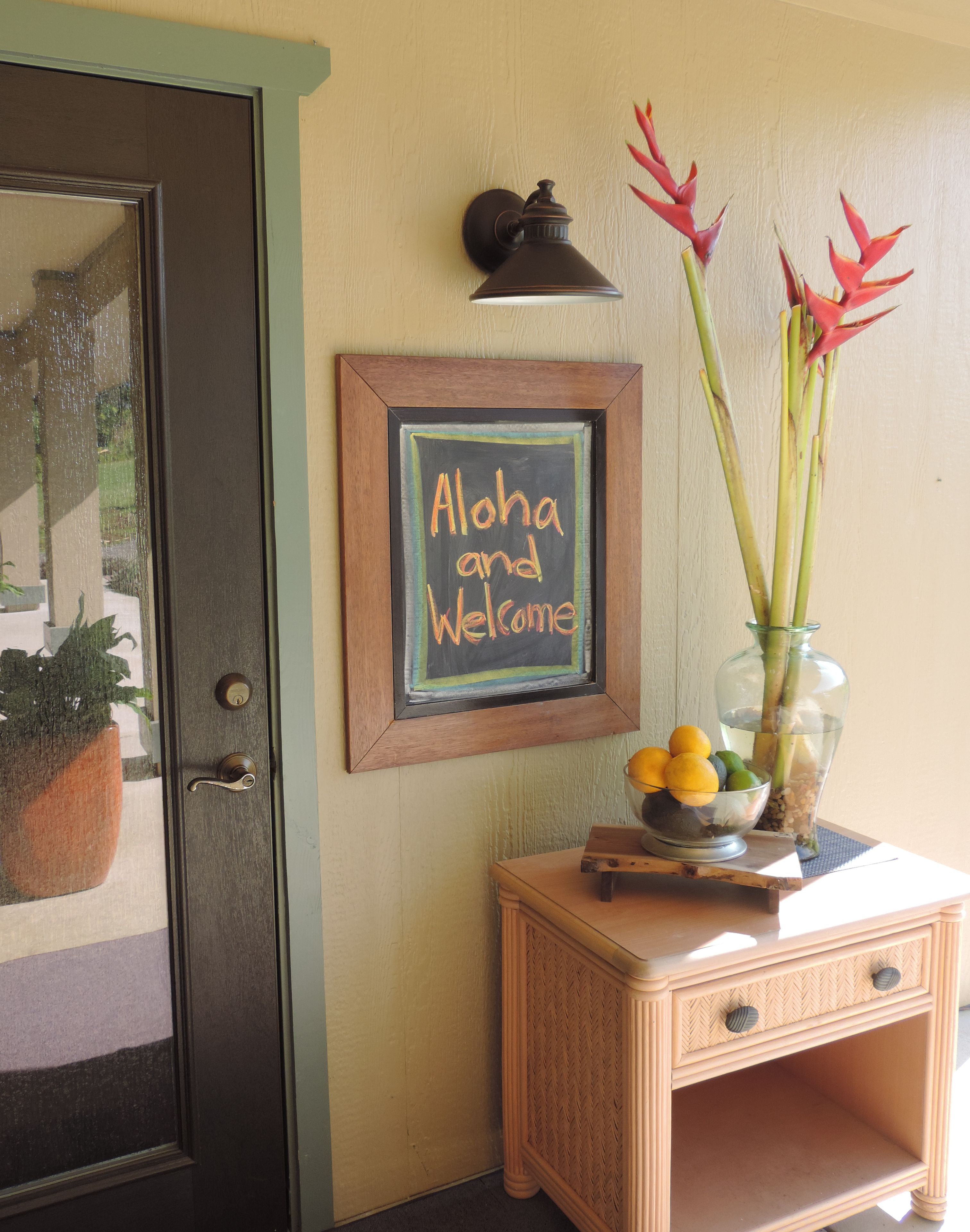 Una puerta con marco oscuro, una lámpara de pared, un cartel enmarcado que dice 'Aloha and Welcome', y una mesa pequeña con un jarrón de flores y limones.