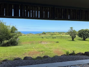 A view from a covered area to a grassy field with trees and a distant view of the ocean.