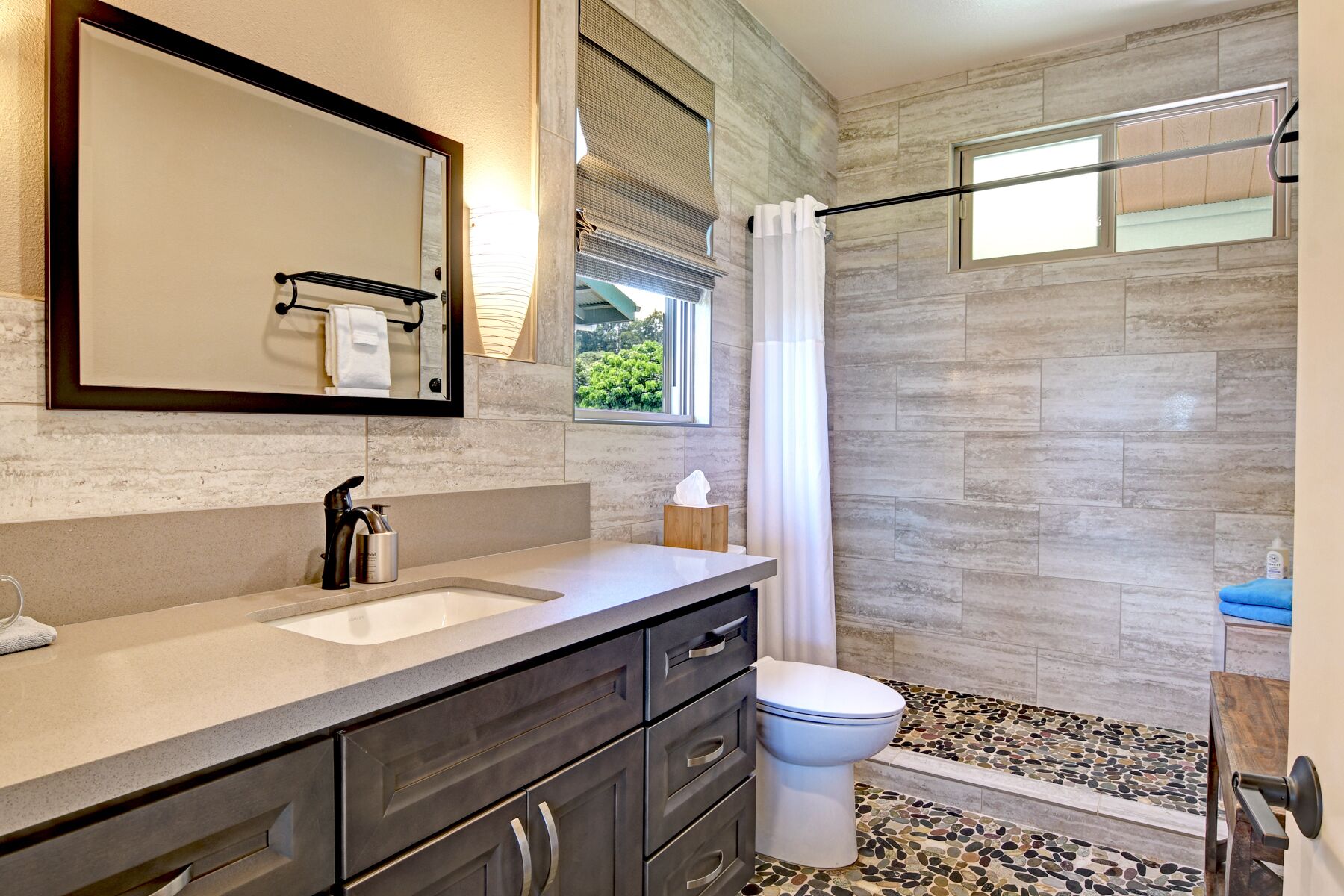 A modern bathroom with a large mirror, a sink, a toilet, and a shower area with a pebble floor.