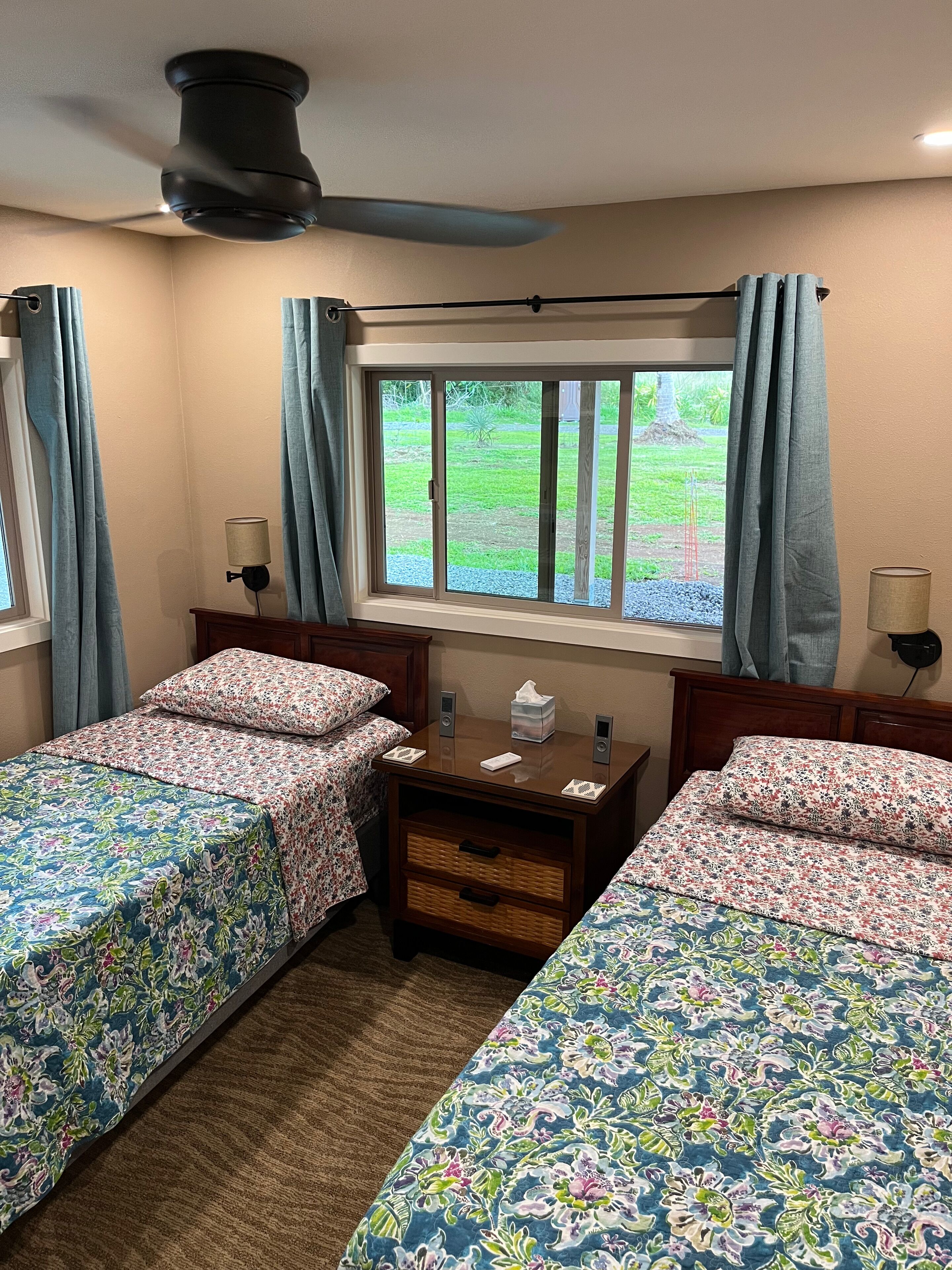 A room with two beds, a window with curtains, a bedside table with a lamp, and a ceiling fan.