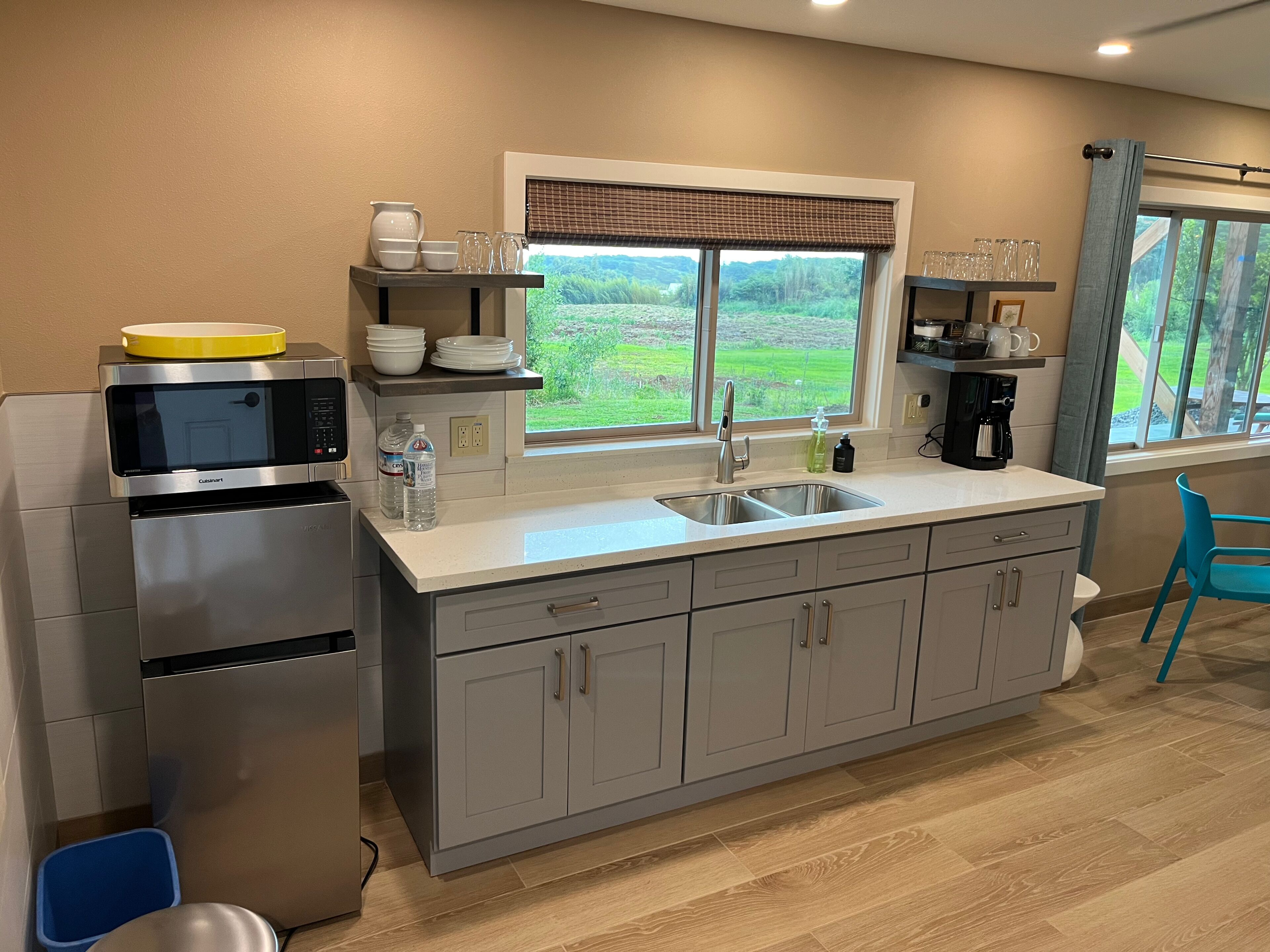 A modern kitchen with a built-in microwave, stainless steel appliances, a large window with a view of greenery, and a blue chair.