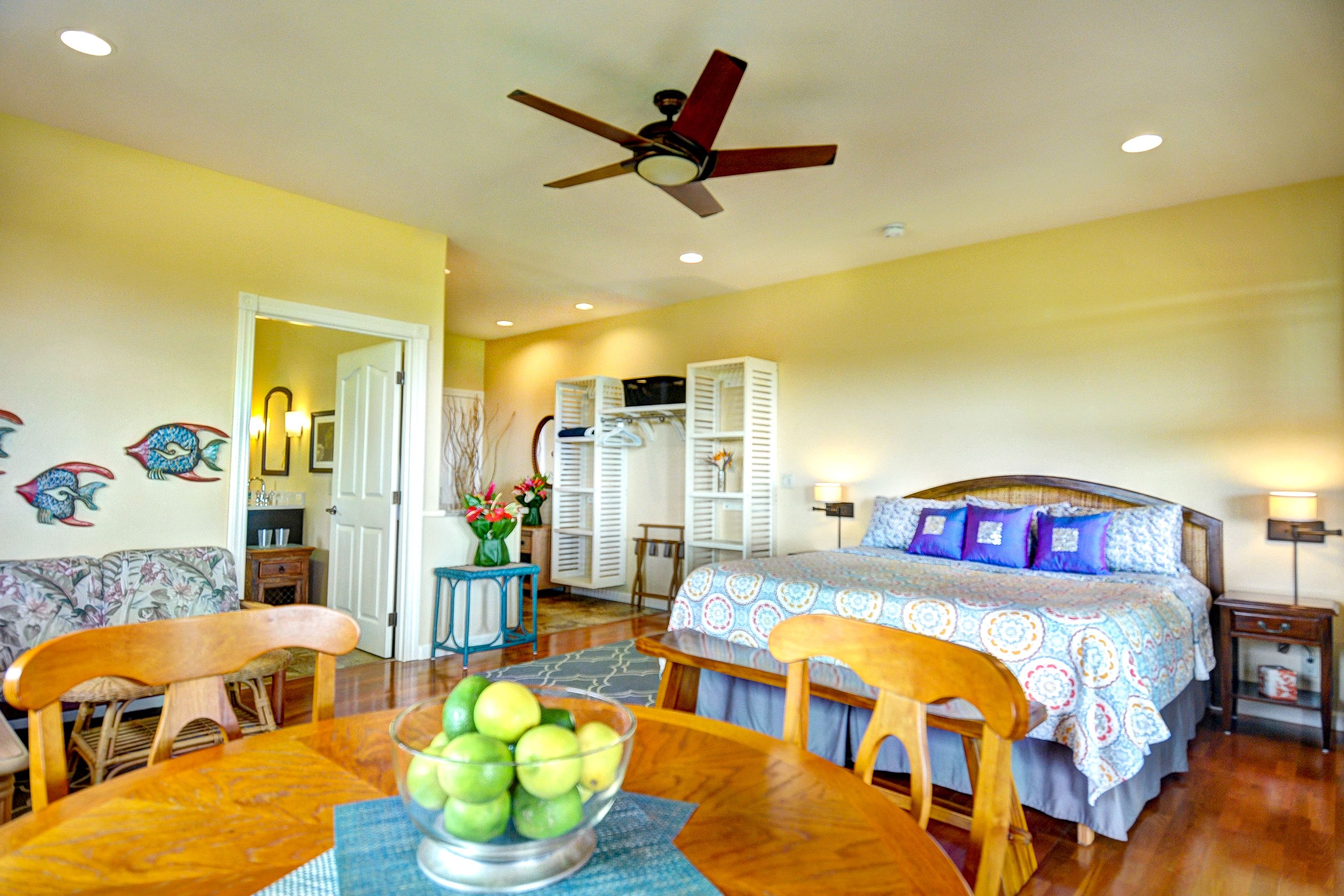 A room with a bed, a dining table with chairs, a ceiling fan, and a bowl of fruit.