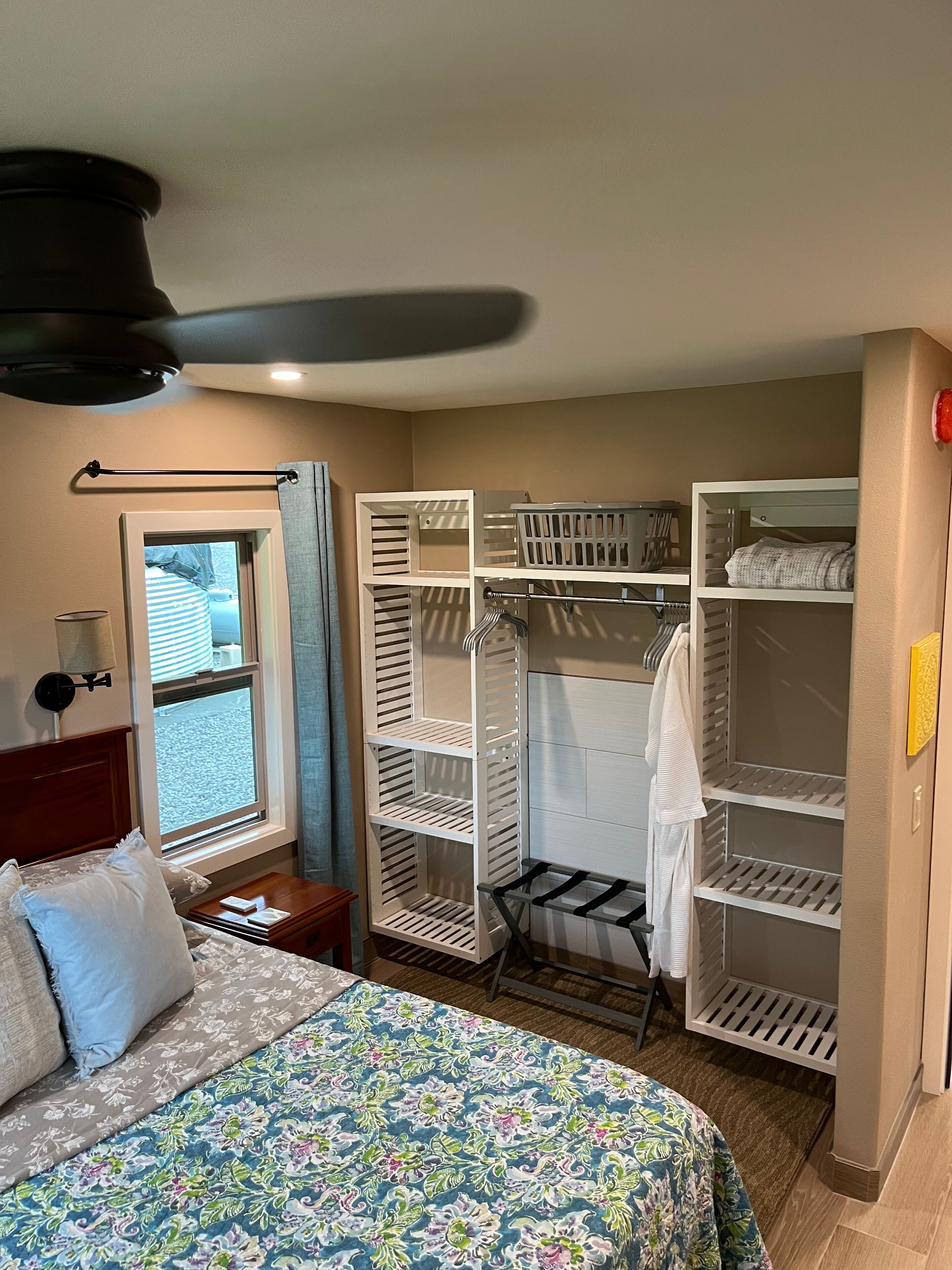 A hotel room with a bed, a window, a ceiling fan, and a walk-in closet with shelves.