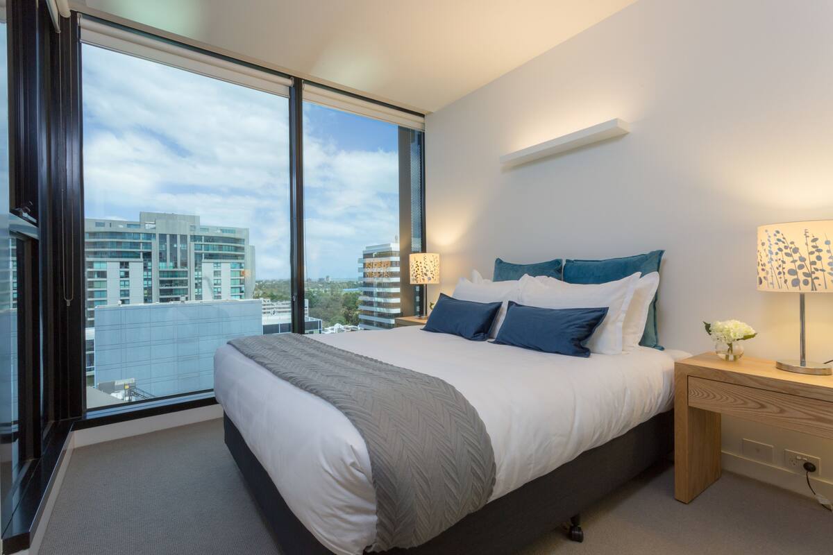 A modern hotel room with a large bed, a nightstand, a lamp, and a view of the city through large windows.