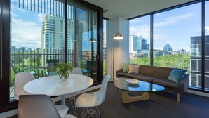 Een moderne woonkamer met een glazen tafel, witte stoelen, een bank met kussens en grote ramen met uitzicht op de stad.
