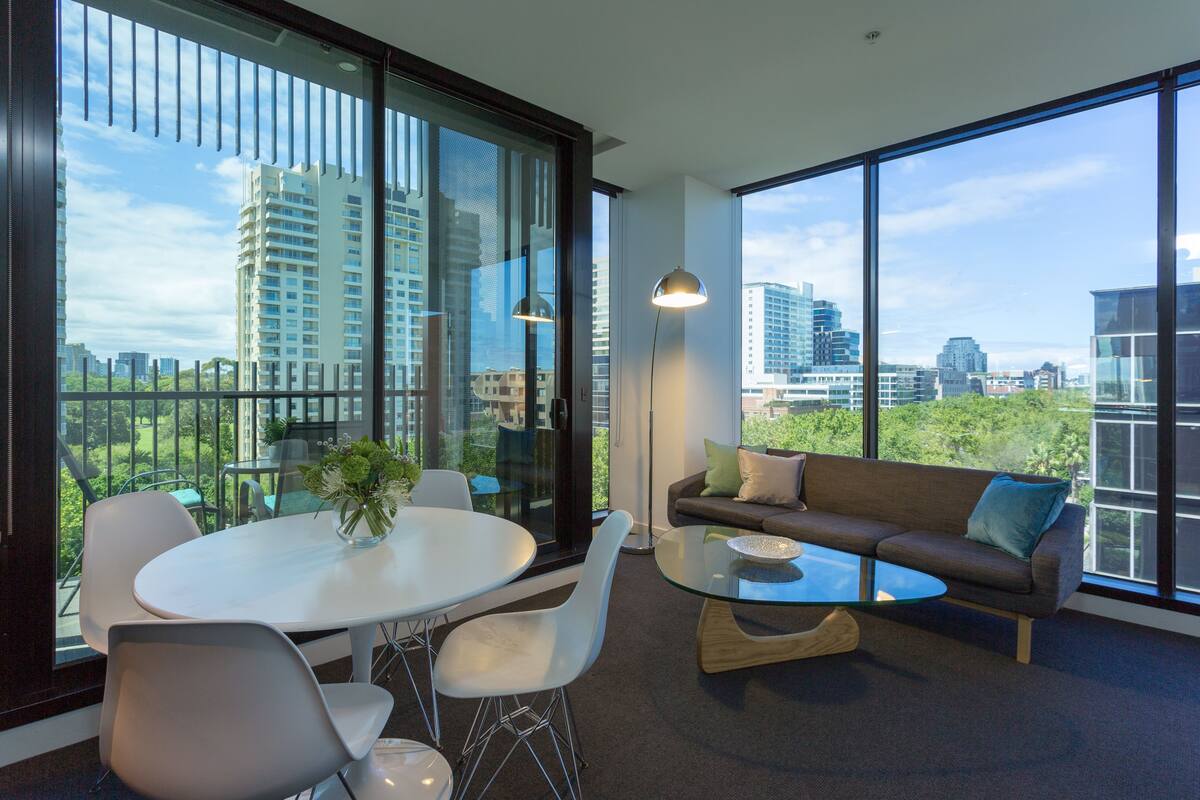 A modern living room with a glass table, white chairs, a sofa with cushions, and large windows offering a city view.