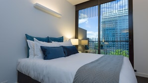 Een moderne hotelkamer met een groot bed, een nachtkastje, een lamp en uitzicht op een stadslandschap door een groot raam.