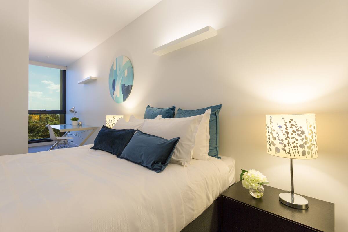 A modern bedroom with a large bed, a bedside table with a lamp, and a view of the outdoors through a window.
