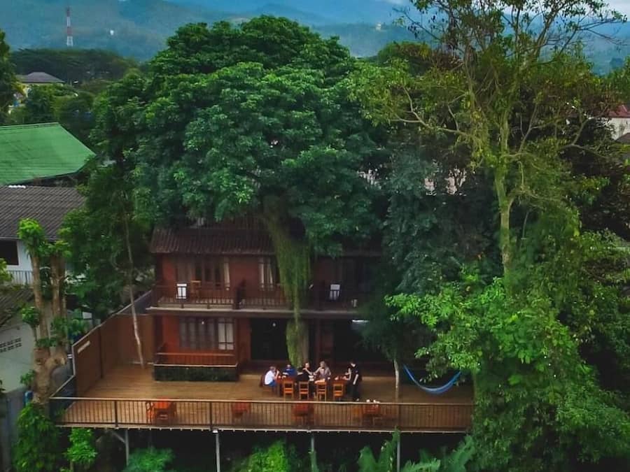 Una casa con terraza de madera y balcón con vistas a un jardín exuberante y montañas.