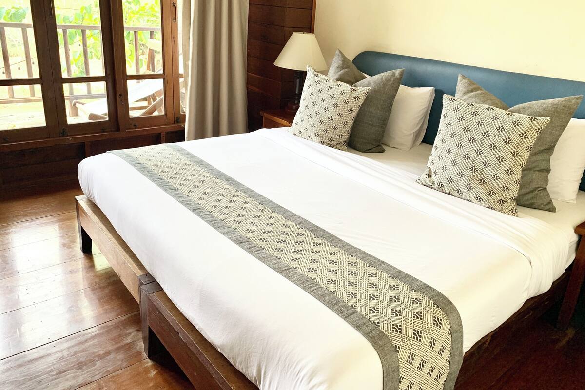 Una cama grande con un corredor estampado, varios almohadones, un cabecero de madera y una mesita de noche con una lámpara.