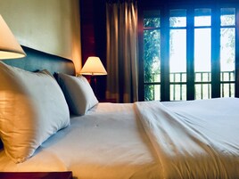 Una cama bien hecha con sábanas y almohadas blancas, una lámpara de noche y una ventana con cortinas.