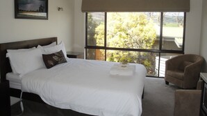 A hotel room with a large bed, a chair, a small table, and a window with a view of trees. - Delatite Apartments Merrijig (Merrijig)