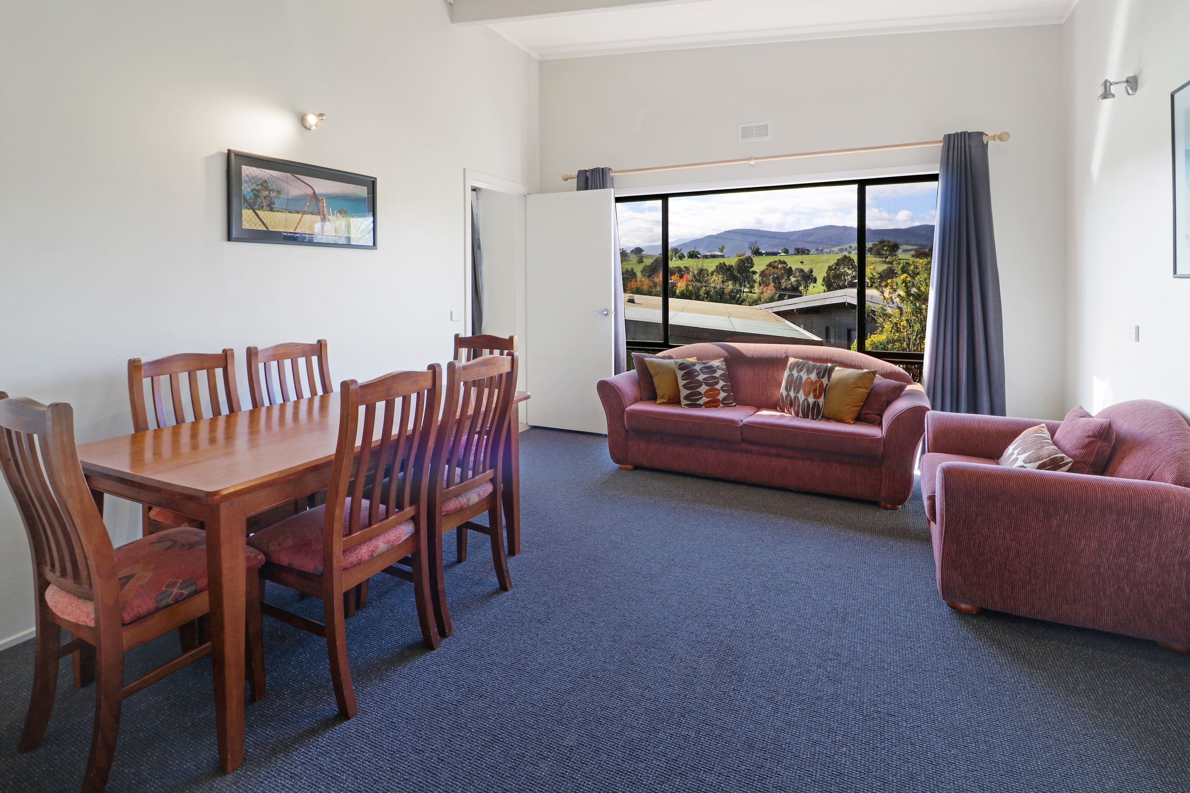 A room with a dining table and chairs, a red sofa, and a large window with a view of the countryside.