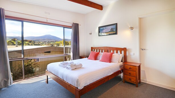 A bedroom with a large bed, a wooden nightstand, and a view of a mountainous landscape through large windows. - Delatite Apartments Merrijig (Merrijig)