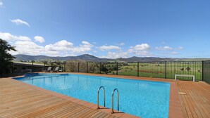 A swimming pool with a wooden deck and metal railings, surrounded by a fence and a view of distant hills. - Delatite Apartments Merrijig (Merrijig)