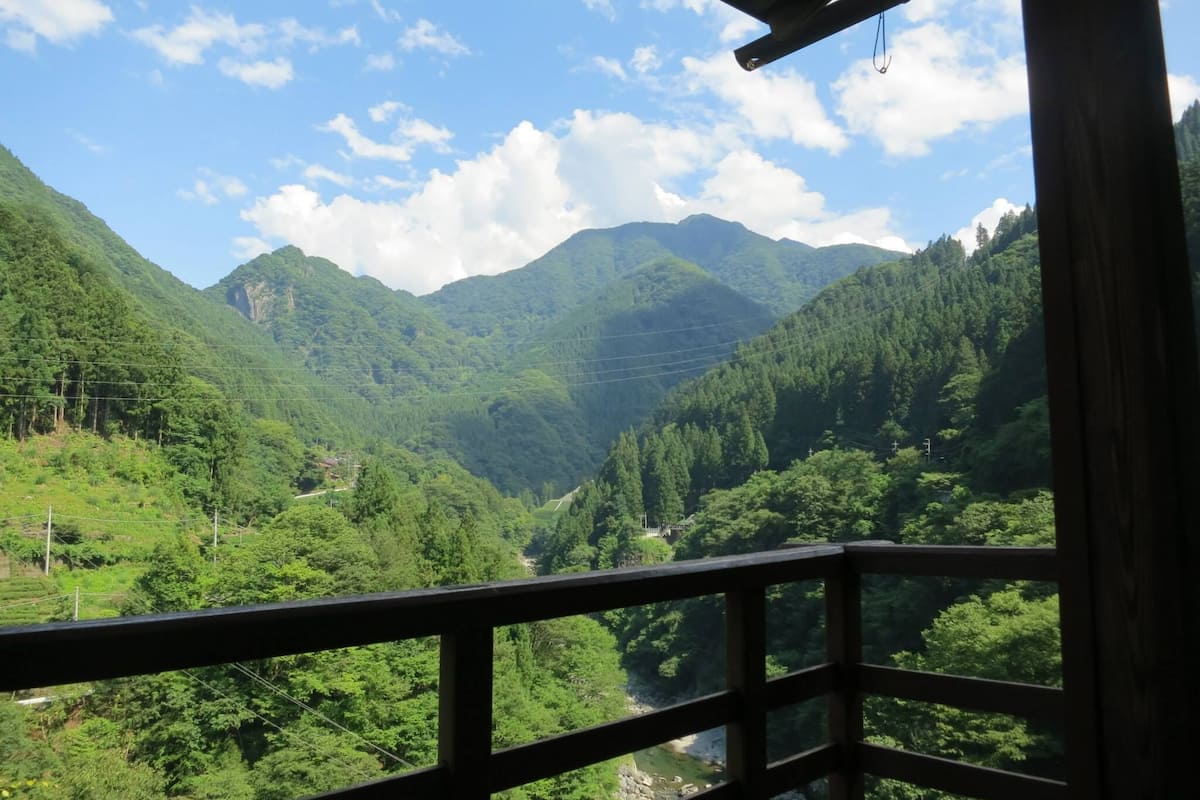 Une vue depuis un balcon doté d’une balustrade en bois, donnant sur une vallée verdoyante, parsemée d’une végétation luxuriante, et sur des montagnes lointaines.