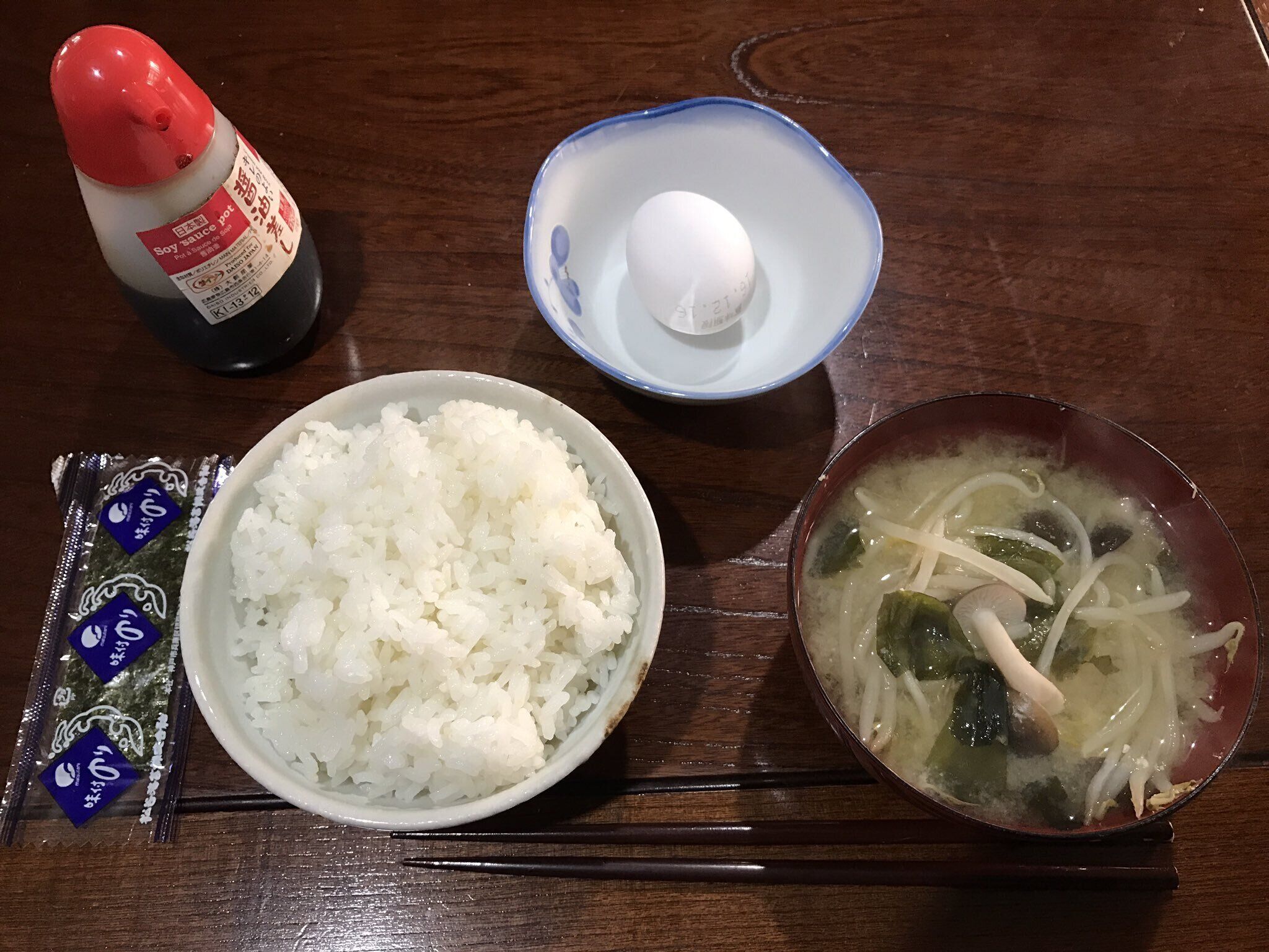 A table with a bowl of white rice, a bowl of soup with vegetables, a packet of seaweed, and a bottle of soy sauce.