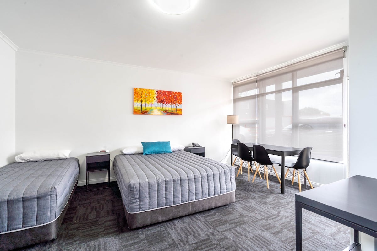 A hotel room with two single beds, a desk, a chair, a table, and a large window with blinds.