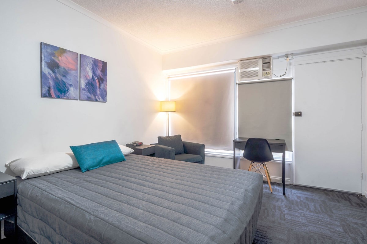 A hotel room with a large bed, a desk, a chair, a lamp, and a window.