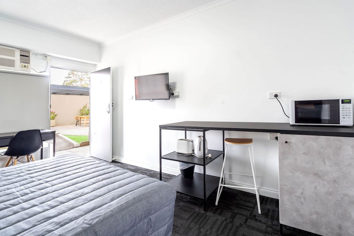 A hotel room with a bed, a desk with a computer, a TV, a microwave, and a small stool.