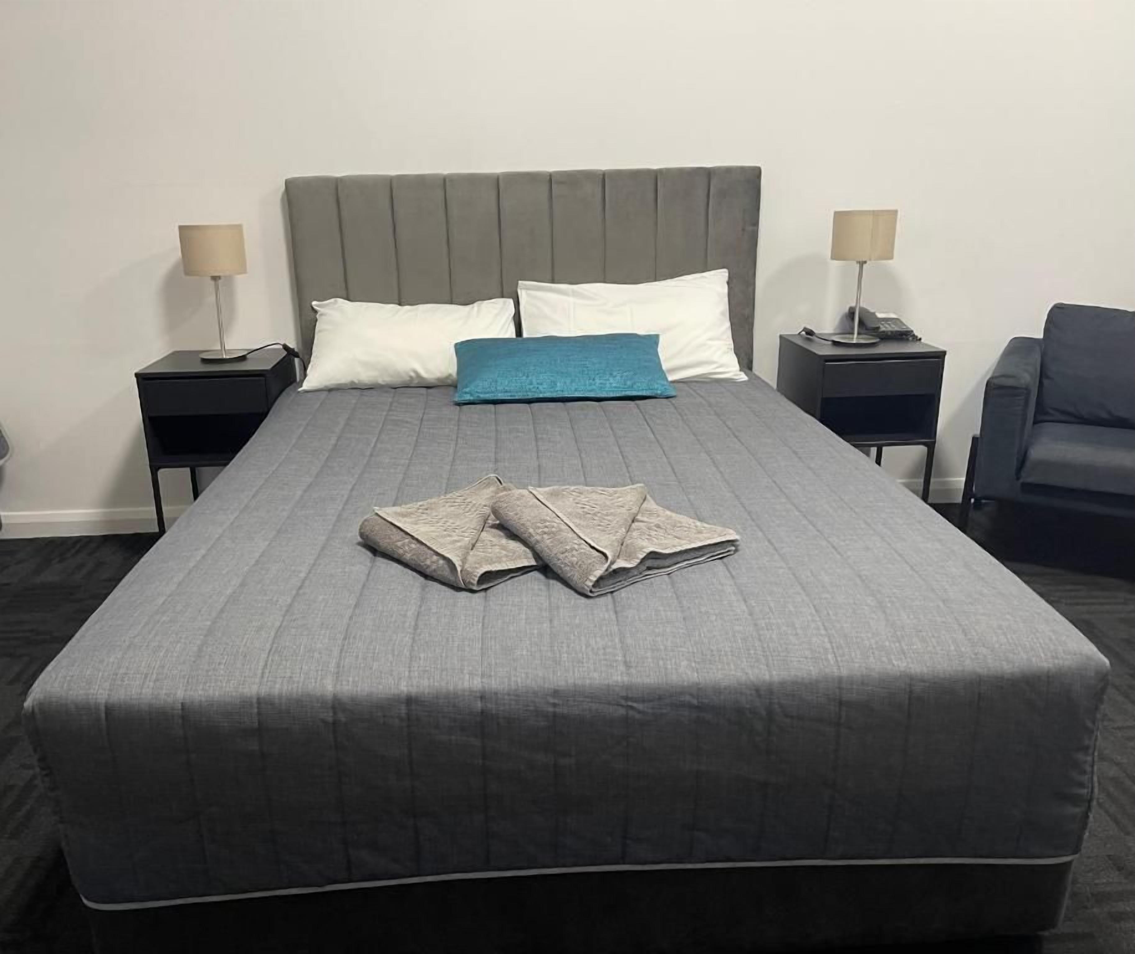 A neatly made bed with a grey bedspread, two white pillows, and a blue accent pillow. There are two bedside tables with lamps, and a dark-colored armchair to the right.
