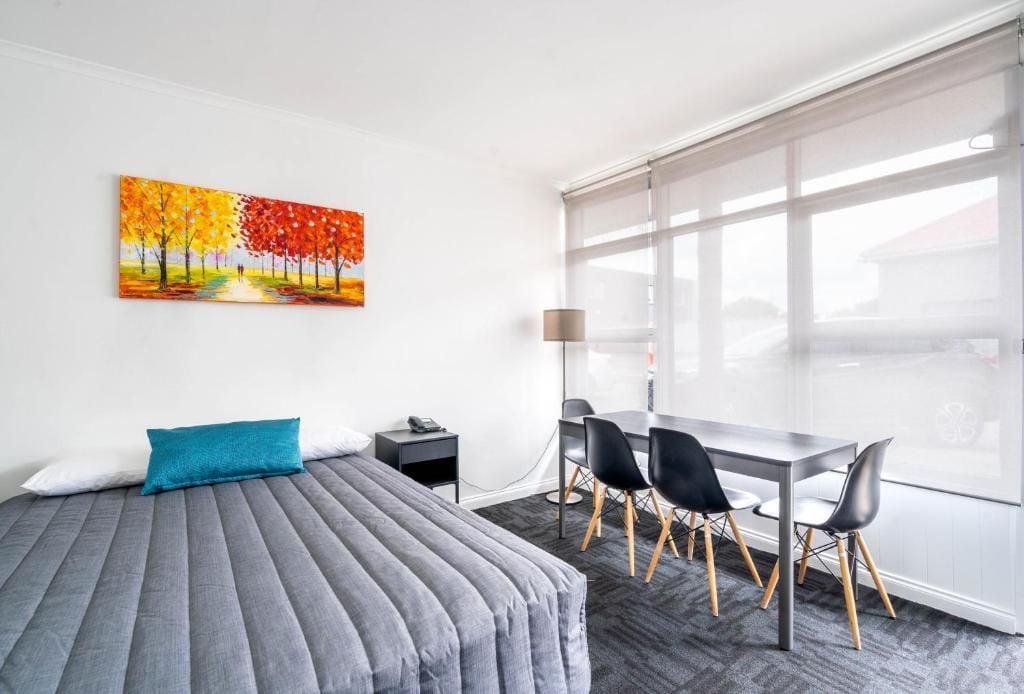 A hotel room with a bed, a desk, and chairs, a painting on the wall, and a large window.