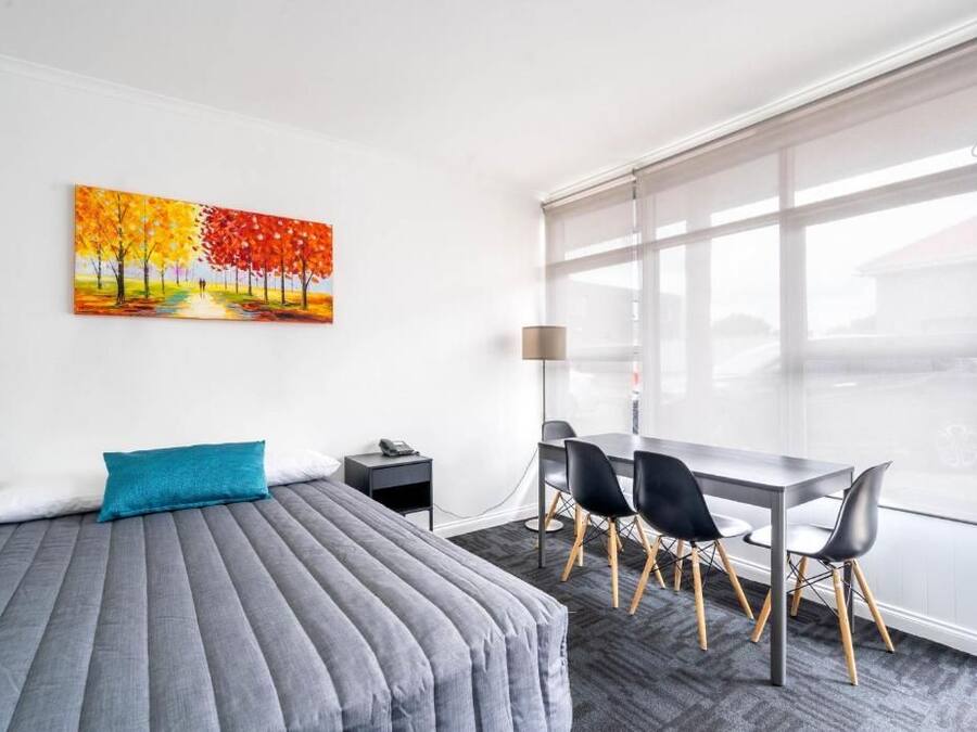 A hotel room with a bed, a desk, and chairs, a painting on the wall, and a large window.