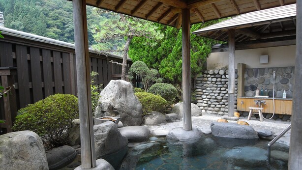 Un onsen japonés tradicional con una terraza de madera, rocas grandes y un muro de piedra.