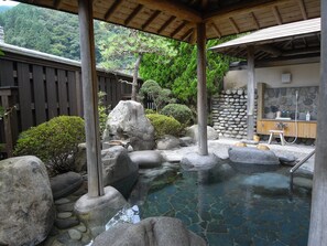Un onsen japonés tradicional con una terraza de madera, rocas grandes y un muro de piedra.