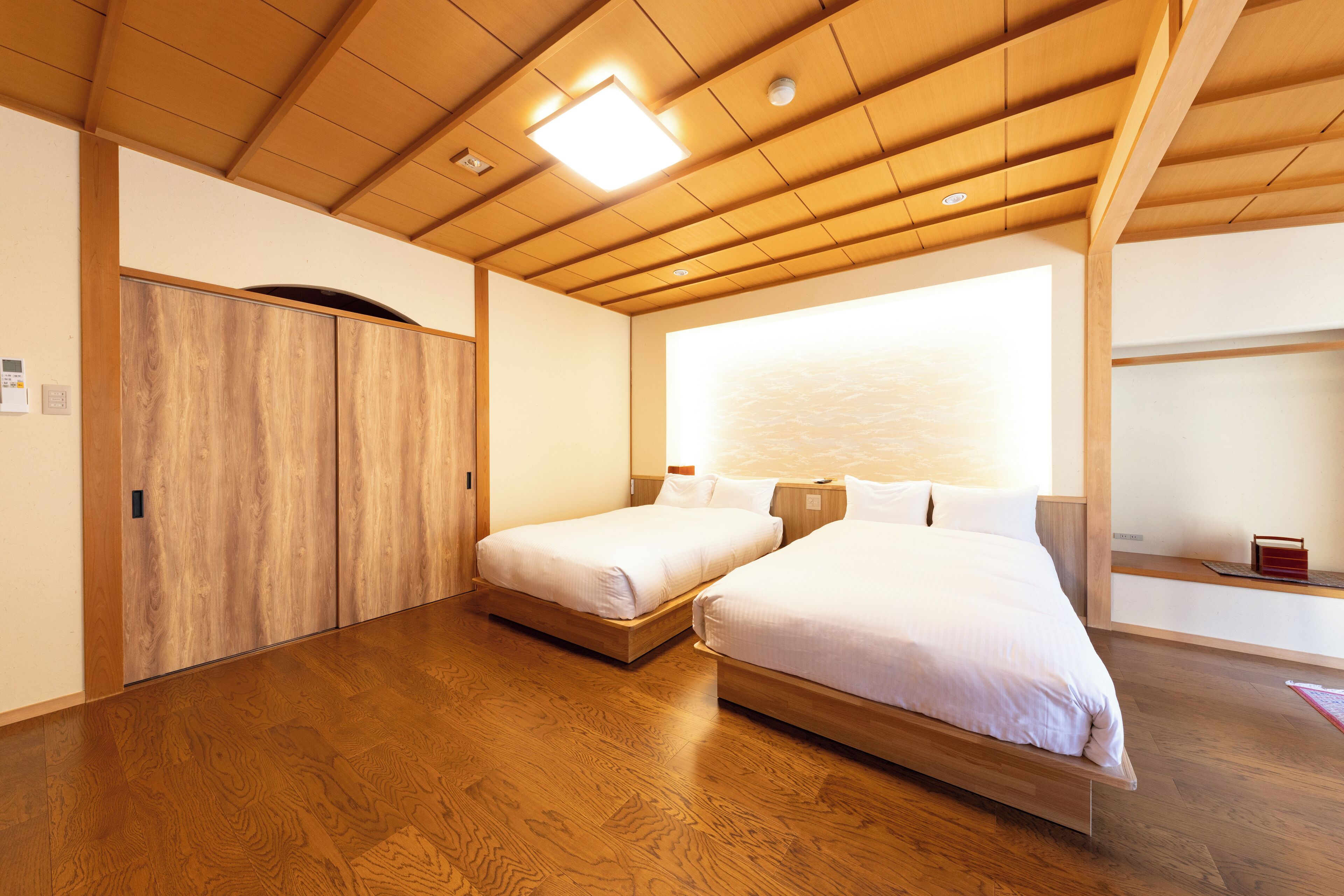 Una habitación con dos camas, suelo de madera, un ventanal grande y un techo con vigas de madera e iluminación empotrada.