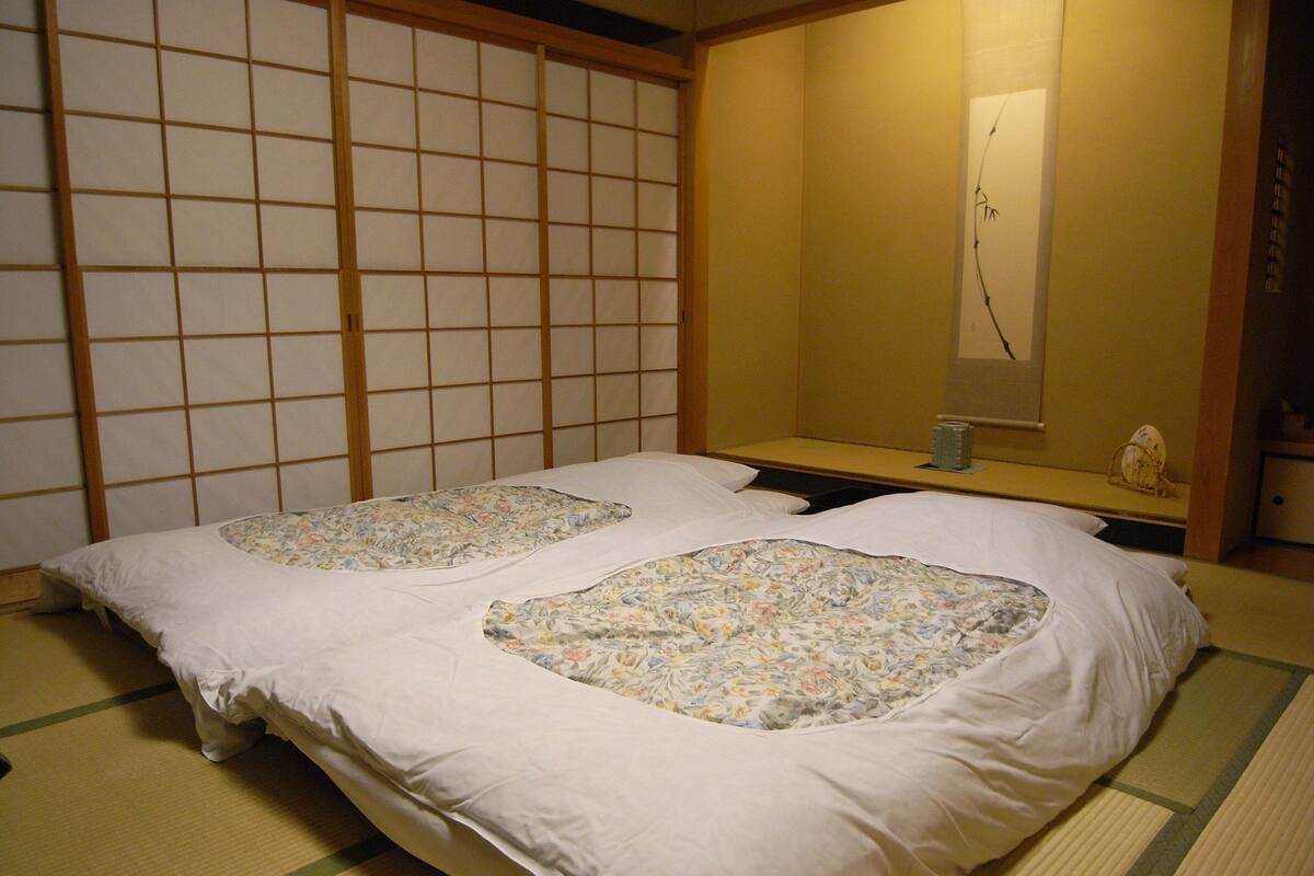Ein traditioneller japanischer Raum mit Tatami-Boden, einem niedrigen Bett mit floralem Bettbezug und einer raumteilenden Shoji-Schiebetür.