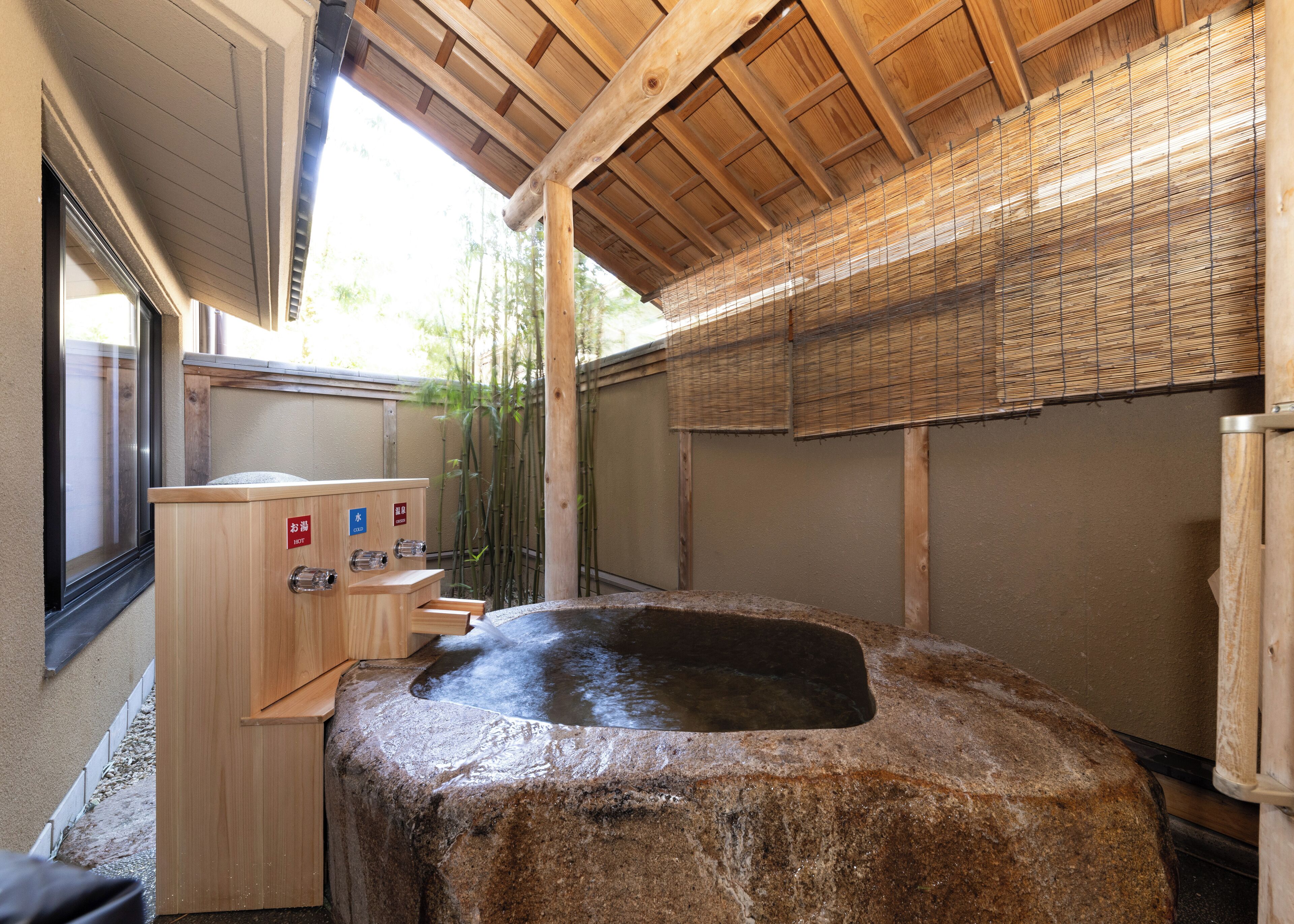 Un onsen japonés tradicional con una fuente termal de piedra, bancos de madera y biombos de bambú.