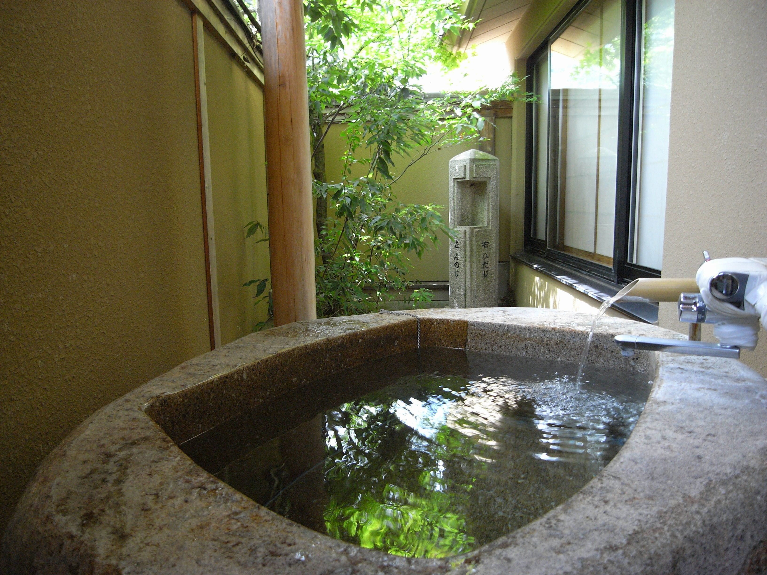Een stenen hottub met stromend water, omringd door een gebouw met een raam en een boom.