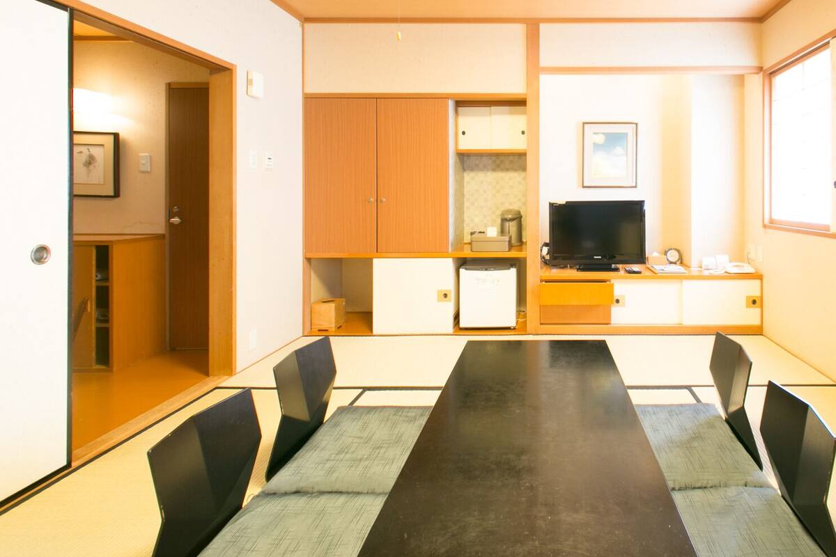 A compact room with a dining table, chairs, a television, and a small kitchenette.