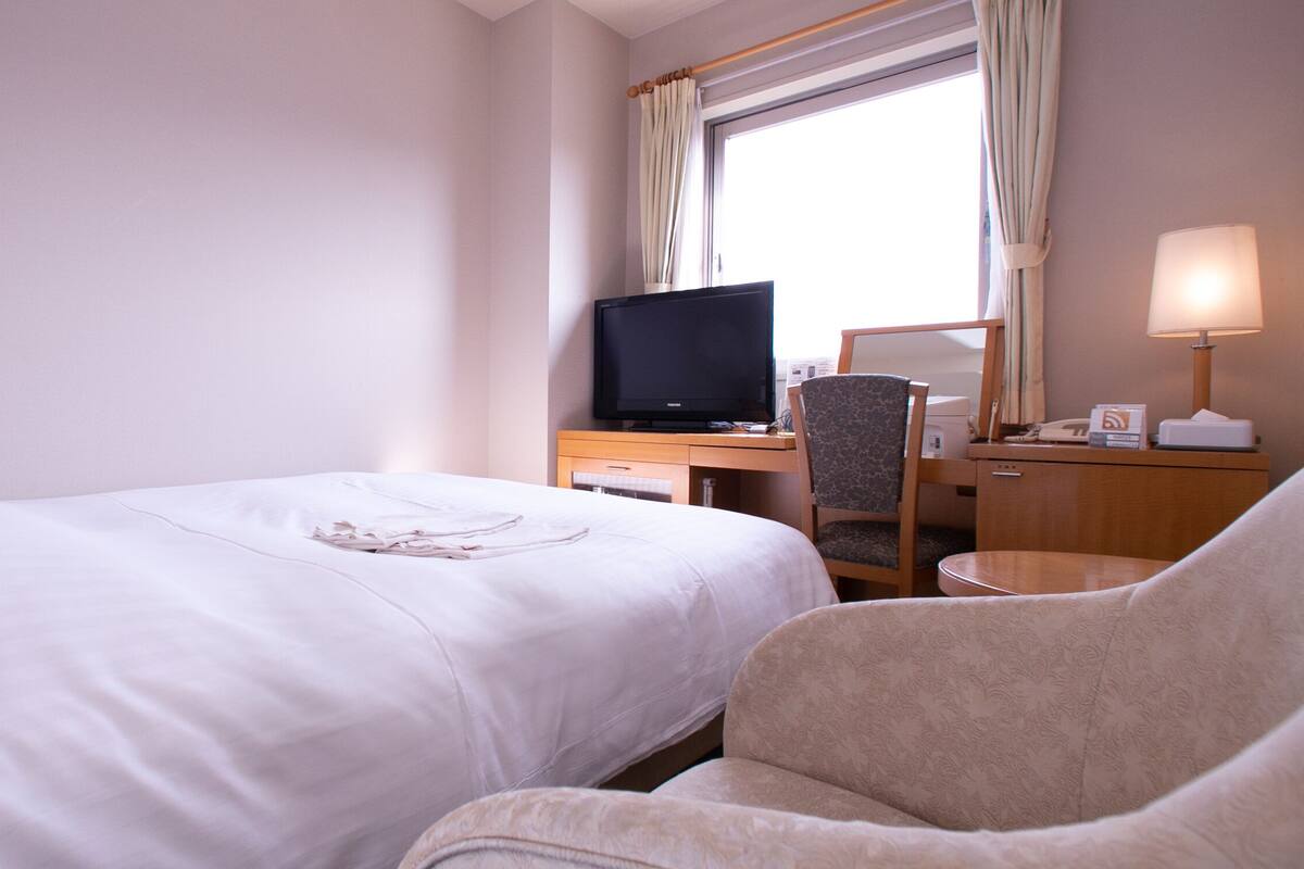 A hotel room with a bed, a desk with a television, a chair, and a lamp.