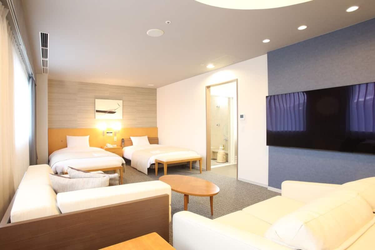 A hotel room with two beds, a sofa, a small table, a TV, and a bathroom door.