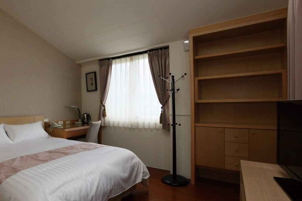 Quarto de hotel com cama, escrivaninha com luminária, cabideiro, televisão e armário.