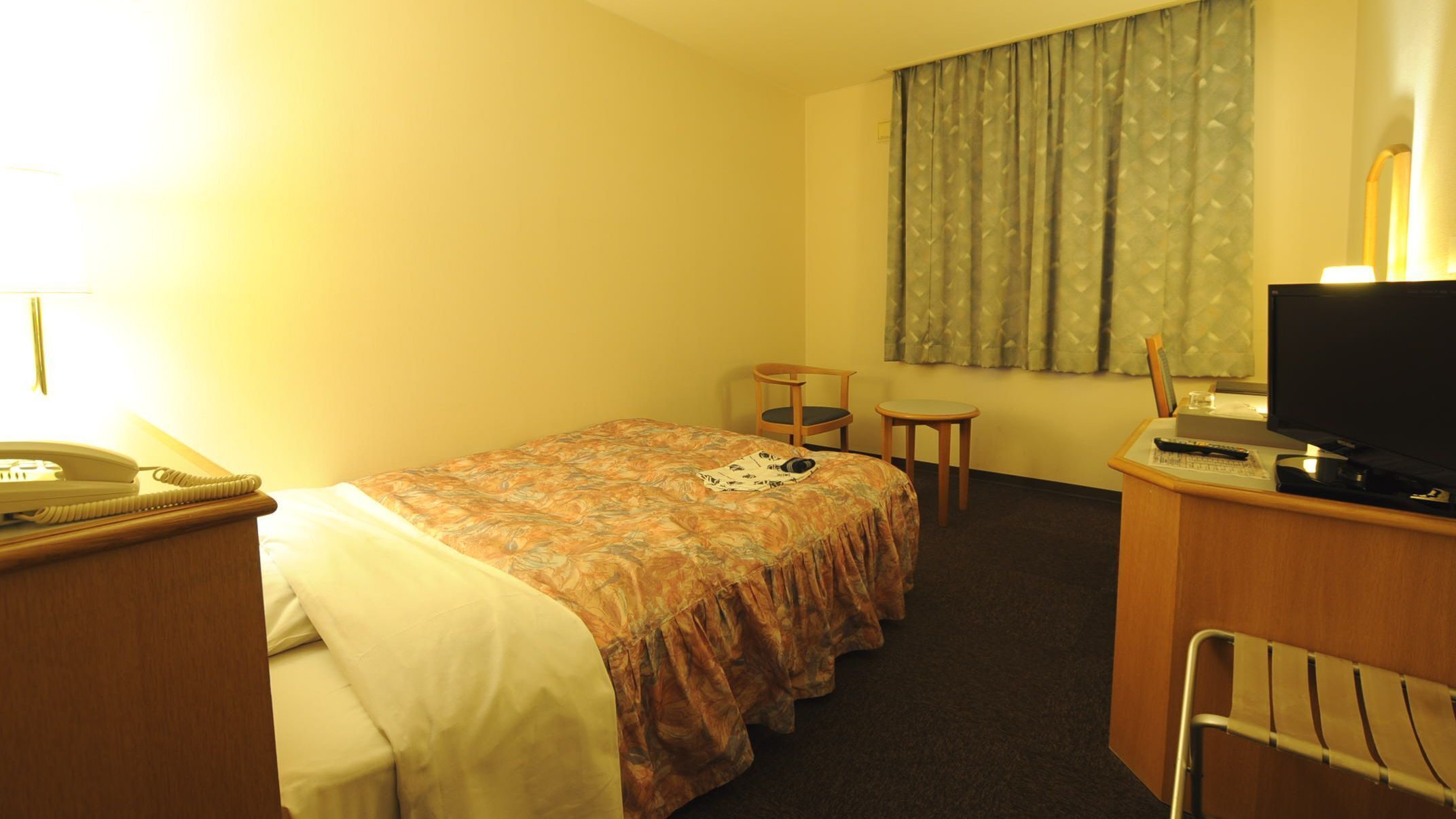 Una habitación de hotel con una cama, un escritorio con computadora, una silla, teléfono y una ventana con cortinas.