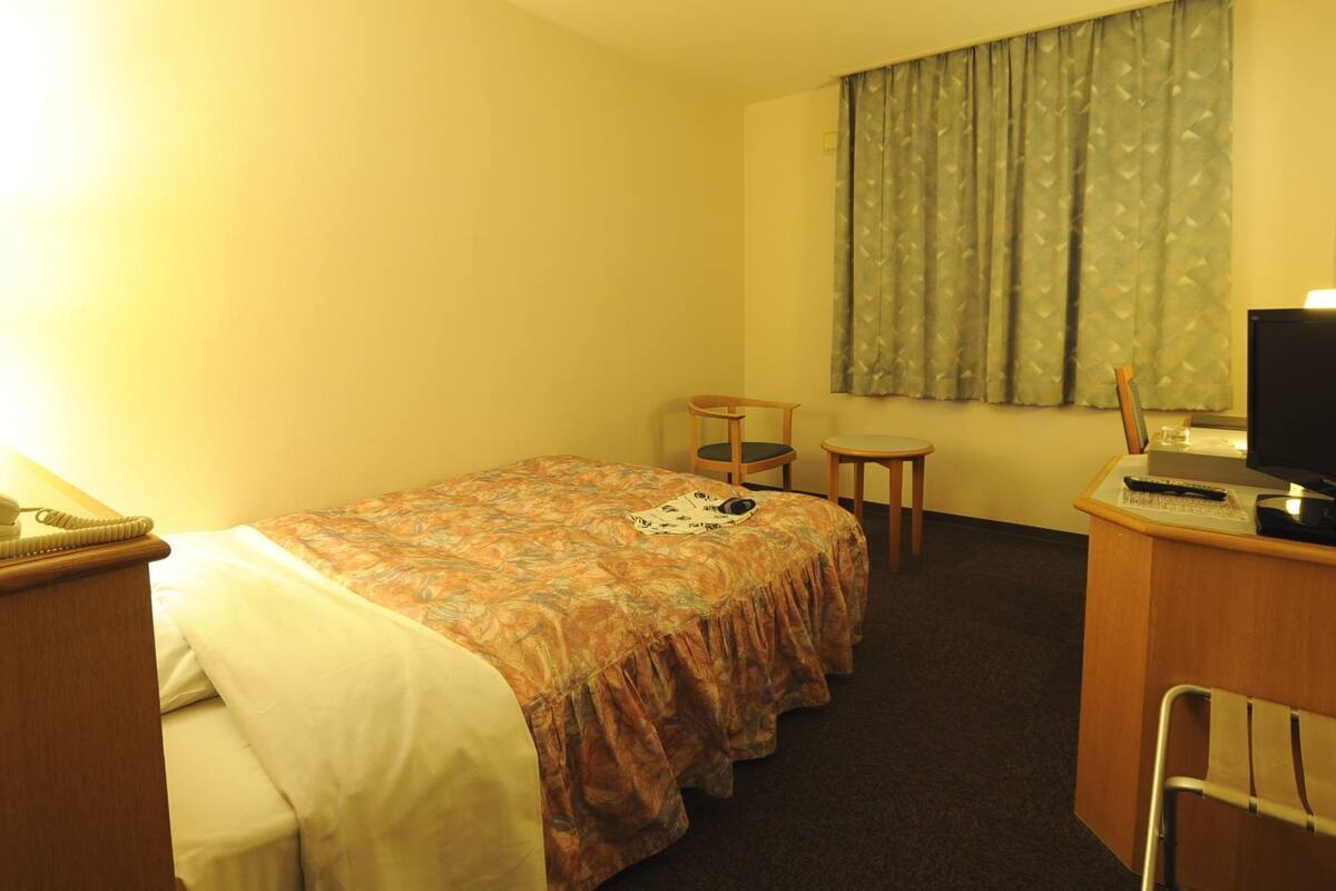 Ett hotellrum med en säng, ett skrivbord med en dator, en stol, en telefon och ett fönster med gardiner.