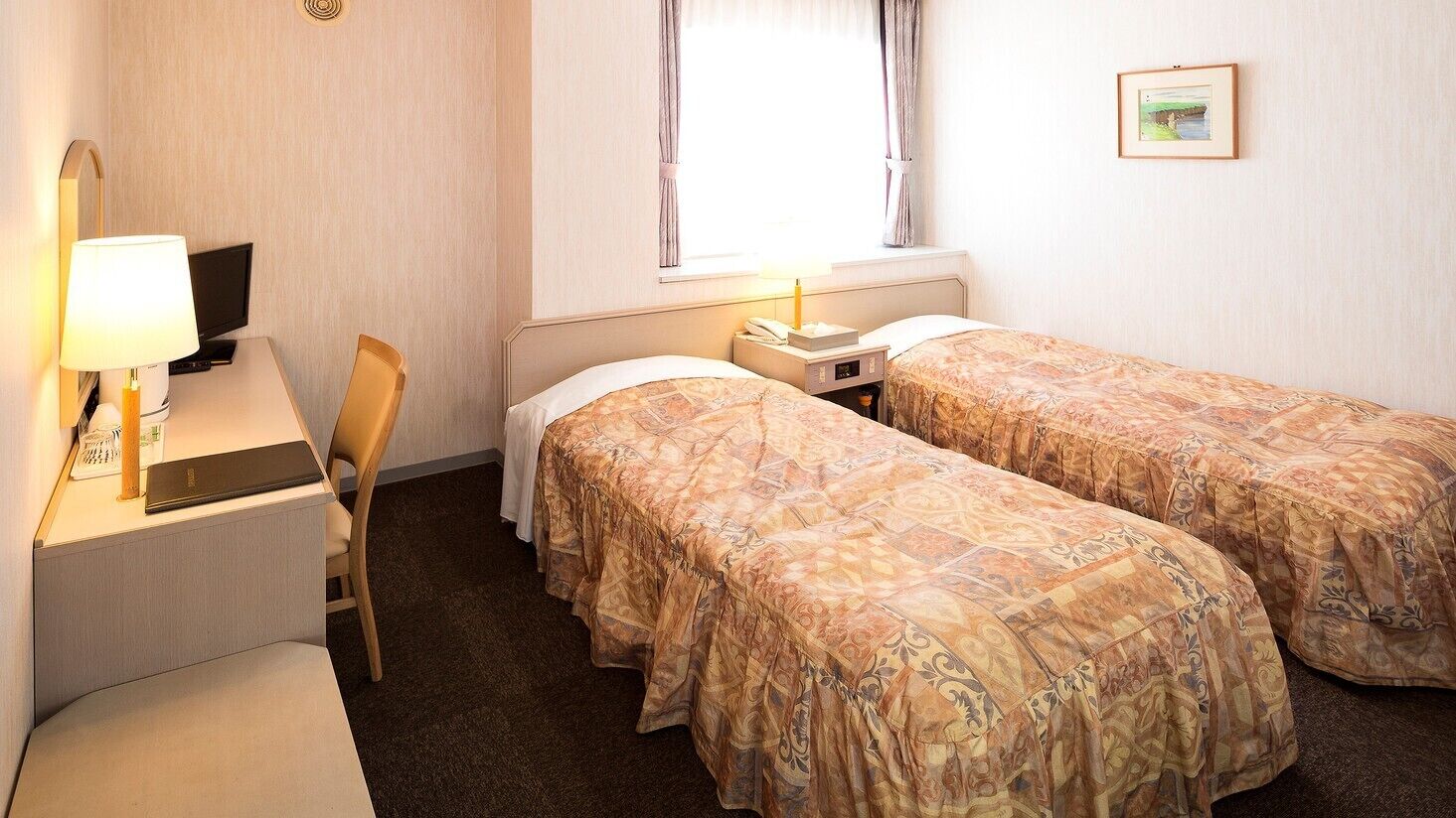 Habitación de hotel con dos camas, un escritorio con computadora, una silla, una mesita de noche con lámpara y una ventana con cortinas.