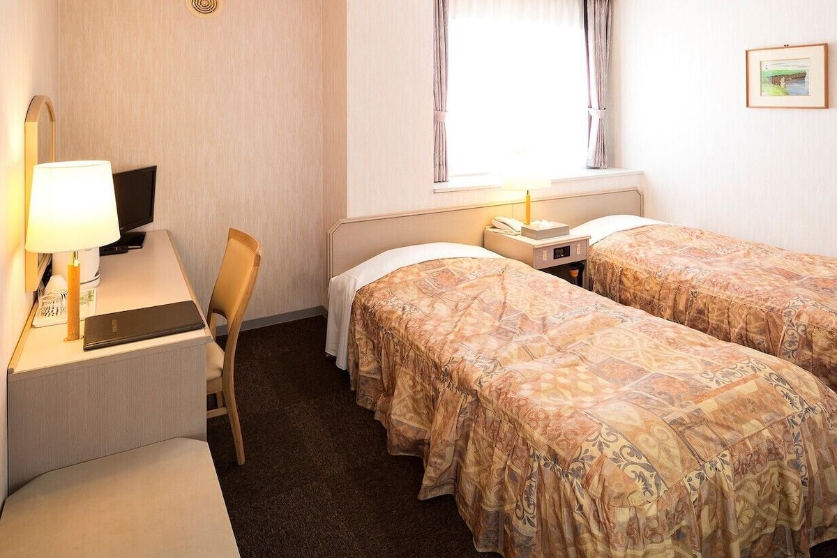 Ett hotellrum med två sängar, ett skrivbord med en dator, en stol, ett nattduksbord med en lampa och ett fönster med gardiner.