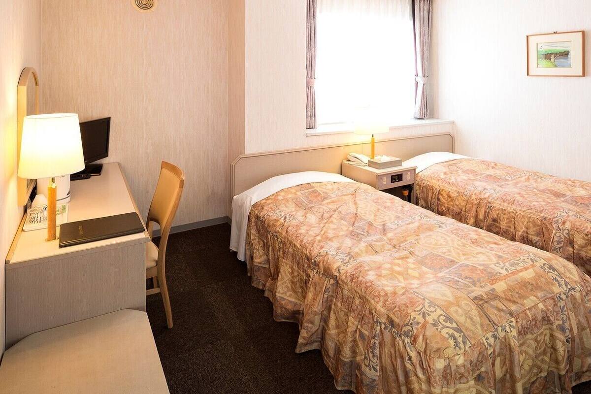 Habitación de hotel con dos camas, un escritorio con computadora, una silla, una mesita de noche con lámpara y una ventana con cortinas.