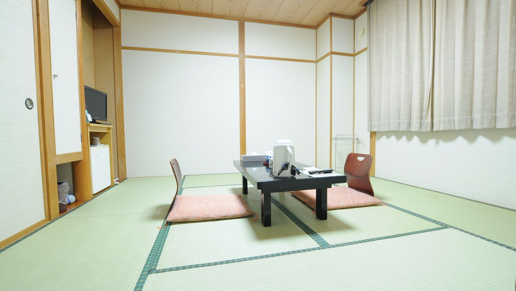 Una habitación japonesa tradicional con suelo de tatami, una mesa baja con un hervidor, una silla y un televisor sobre una estantería.