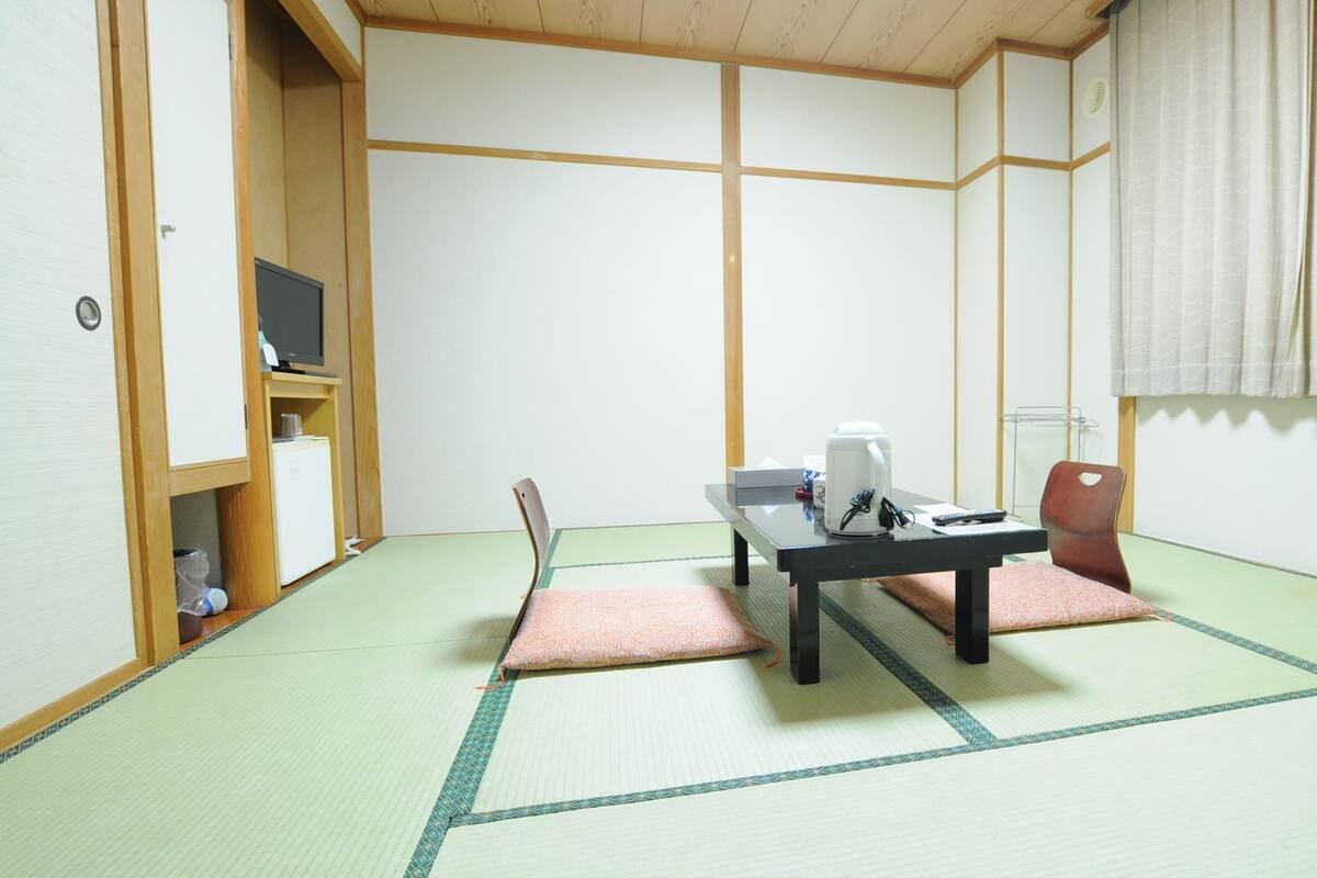 Ett traditionellt japanskt rum med tatamimattor på golvet, ett lågt bord med en vattenkokare, en stol och en tv som står på en hylla.