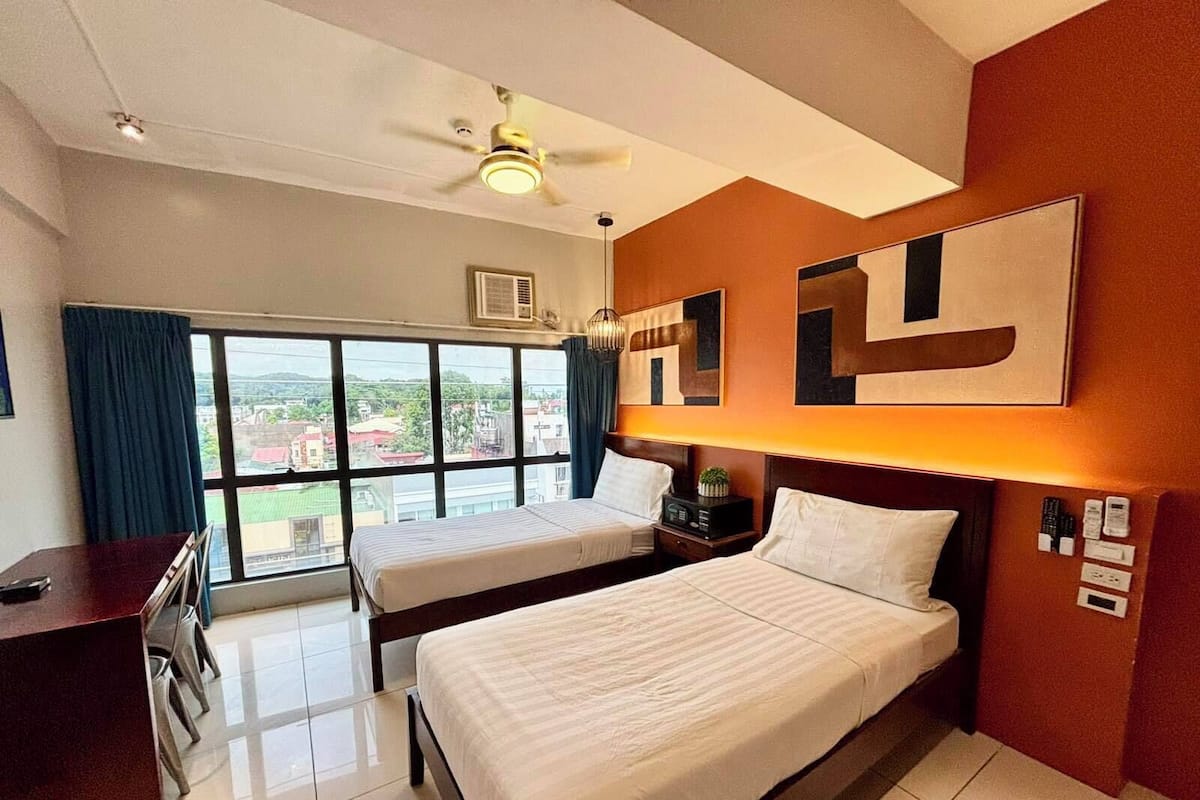 A hotel room with two beds, a desk, a ceiling fan, and a large window with a view.