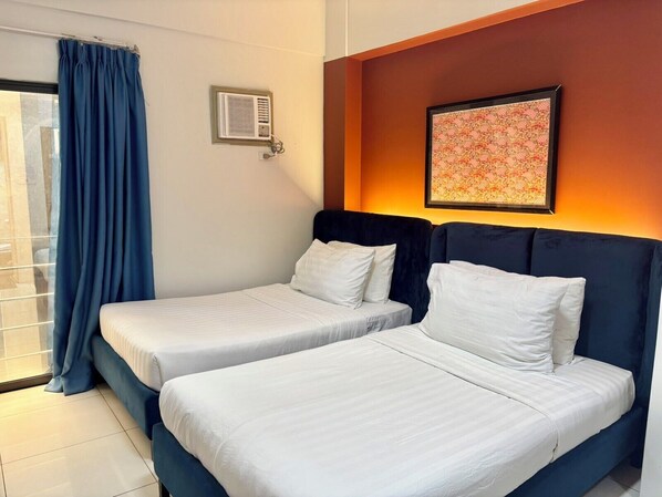 Habitación de hotel con dos camas, una ventana con cortinas, un equipo de aire acondicionado y un cuadro en la pared.