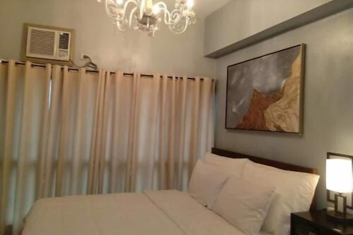 A hotel room with a bed, a chandelier, a painting, a nightstand, and a window with curtains.